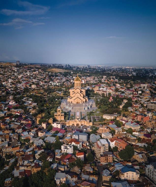 A city seen from above in low sun, a large domed cathedral standing among close-packed rooftops that run away to distant hills.