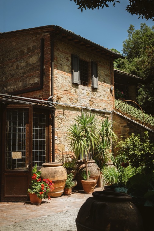 A stone and brick farmhouse with shuttered windows, its courtyard crowded with potted palms, red geraniums and terracotta urns.