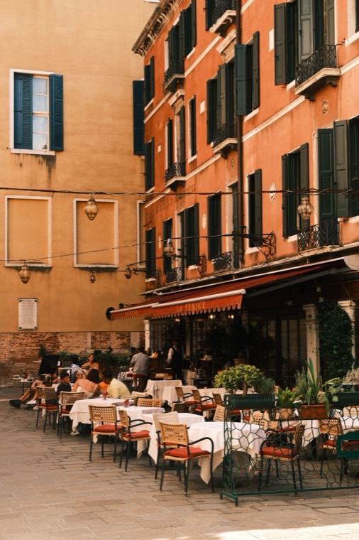 Café tables laid with white cloths and orange cushions along a narrow street, under an awning between ochre and terracotta buildings with green shutters.